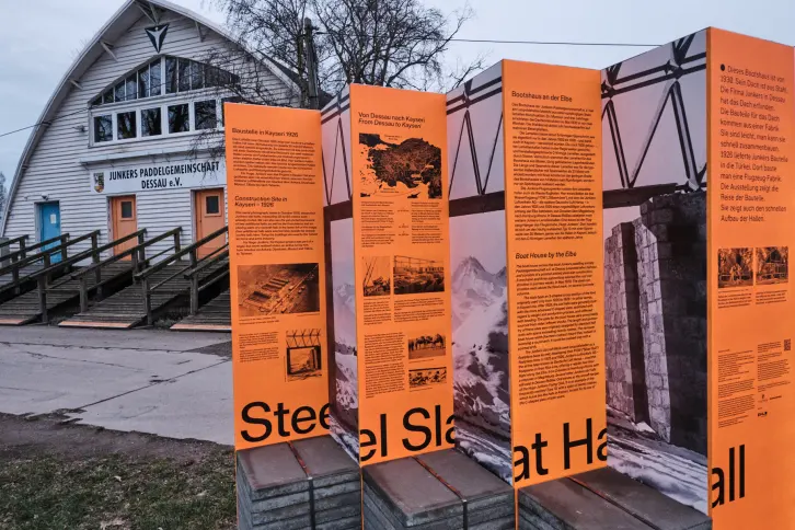 Große Ausstellungstafel vor einem Bootshaus mit Spitzbogen.