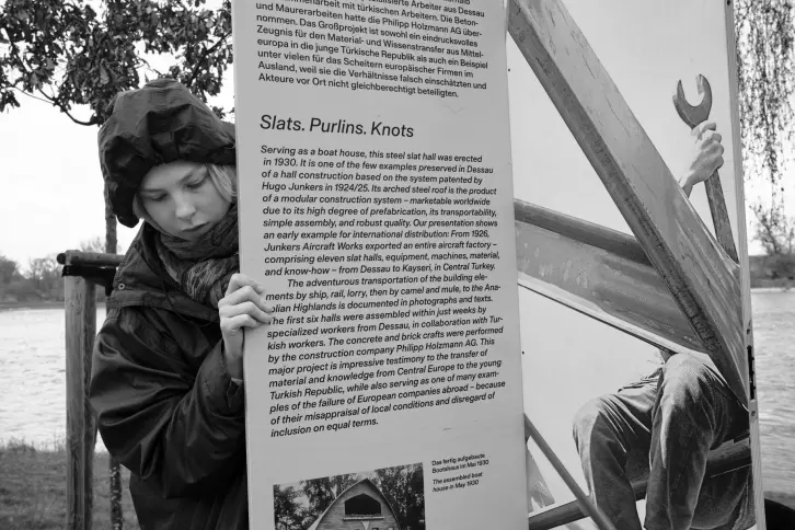 Eine junge Frau in Regenjacke h&auml;lt eine bedruckte Holztafel fest.