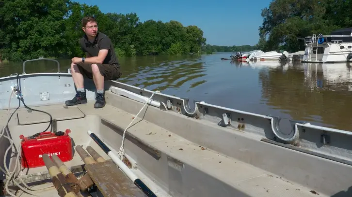 Ein Mann sitzt am Steuer eines Ruderkutters und fährt bei strahlendem Sonnenschein durch einen kleinen, überfluteten Hafen.