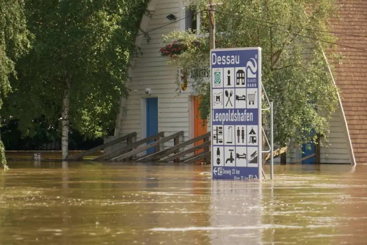Ein überflutetes Bootshaus mit heller Holzfassade und blauen Türen. Schräg ins Wasser führende Bootsrampen ragen aus den Fluten. Eine große Informationstafel weist auf den Leopoldshafen Dessau hin und zeigt Symbole für touristische Einrichtungen. Bäume mit grünem Laub umgeben das Gebäude, Blumen hängen an einem Balkon. Das braune Wasser steht hoch und spiegelt die Umgebung.