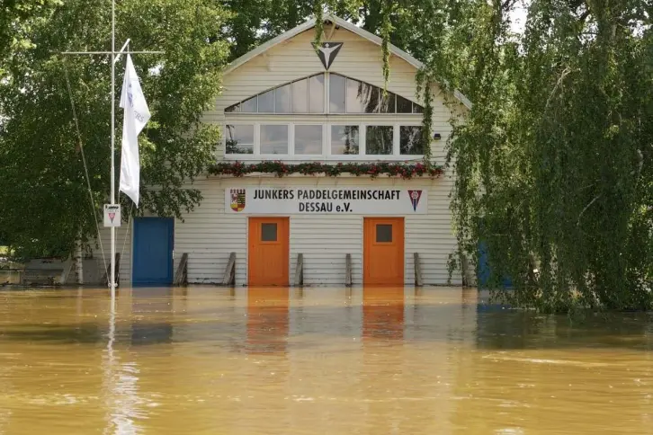 Ein Boothaus mit weißer Holzfassade, mit orangefarbenen und blauen Türen, bis zu denen das bräunliche Hochwasser reicht.