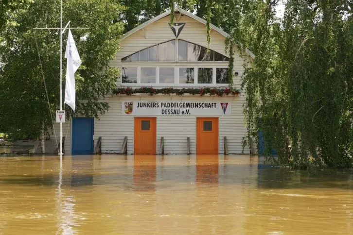 Ein Boothaus mit wei&szlig;er Holzfassade, mit orangefarbenen und blauen T&uuml;ren, bis zu denen das br&auml;unliche Hochwasser reicht.
