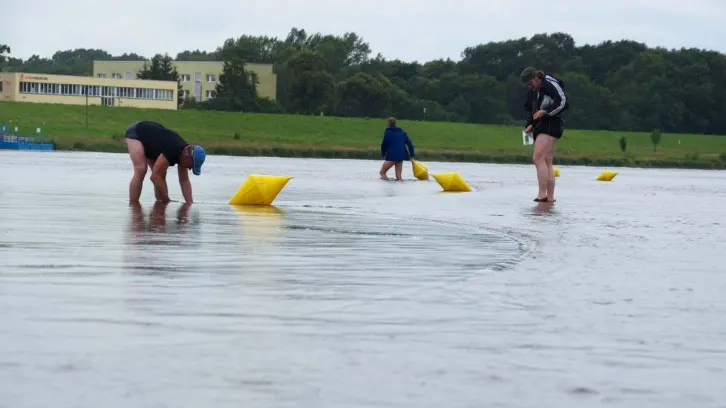 Mehrere Personen stehen im flachen Wasser einer Flusses und setzen gelb Bojen. Im Hintergrund ein Deich, dahinter Gebäude und eine Baumreihe. Der Himmel ist bewölkt.