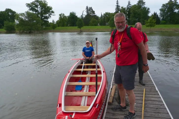 Ein roter 10er-Canadier liegt an einem Holzsteg. Darin sitzt eine Person in blauer Kleidung mit einem Paddel. Am Steg stehen zwei Männer in roten Shirts, einer mit einer Pfeife um den Hals und einem Stechpaddel in der Hand. Der andere trägt Wasserschuhe und hält ein weiteres Paddel. Im Hintergrund ein kleiner Hafen und ein Deich. Der Himmel ist bewölkt.