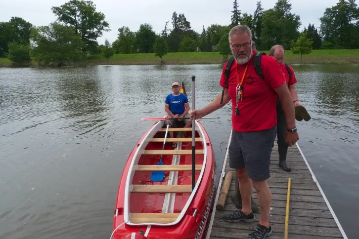 Ein roter 10er-Canadier liegt an einem Holzsteg. Darin sitzt eine Person in blauer Kleidung mit einem Paddel. Am Steg stehen zwei Männer in roten Shirts, einer mit einer Pfeife um den Hals und einem Stechpaddel in der Hand. Der andere trägt Wasserschuhe und hält ein weiteres Paddel. Im Hintergrund ein kleiner Hafen und ein Deich. Der Himmel ist bewölkt.