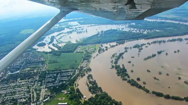 Luftaufnahme einer großflächigen Überschwemmung. Ein brauner Fluss mit über die Ufer getretenem Hochwasser schlängelt sich durch die Landschaft. Teile einer Stadt, landwirtschaftliche Flächen und Wälder sind überflutet. Bäume ragen aus dem Wasser. Der Flügel eines Kleinflugzeugs ist im oberen Bildbereich sichtbar. In der Ferne erstreckt sich die überflutete Flusslandschaft weiter.