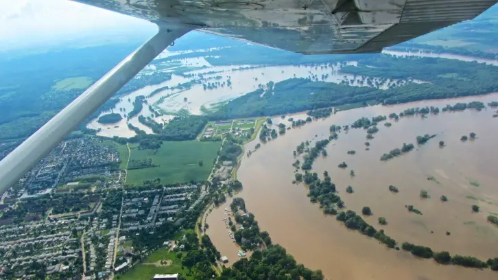 Luftaufnahme einer großflächigen Überschwemmung. Ein brauner Fluss mit über die Ufer getretenem Hochwasser schlängelt sich durch die Landschaft. Teile einer Stadt, landwirtschaftliche Flächen und Wälder sind überflutet. Bäume ragen aus dem Wasser. Der Flügel eines Kleinflugzeugs ist im oberen Bildbereich sichtbar. In der Ferne erstreckt sich die überflutete Flusslandschaft weiter.