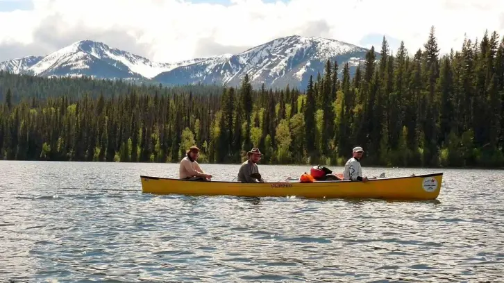 Drei Paddler in einem Canadier auf einem See, im Hintergrund hohe, teils von Schnee bedeckte Berge.