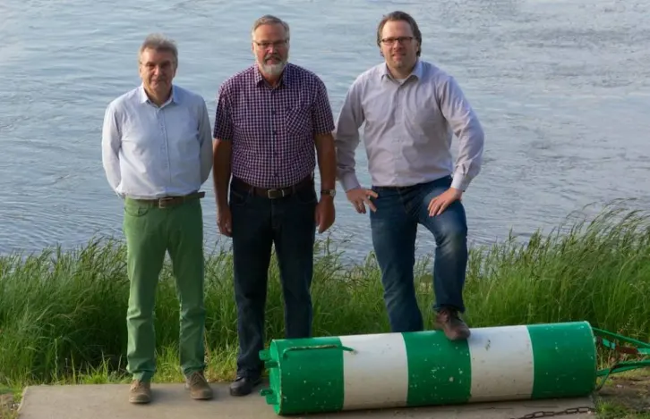 Drei M&auml;nner stehen nebeneinander am Ufer eines Flusses und blicken zur Kamera.