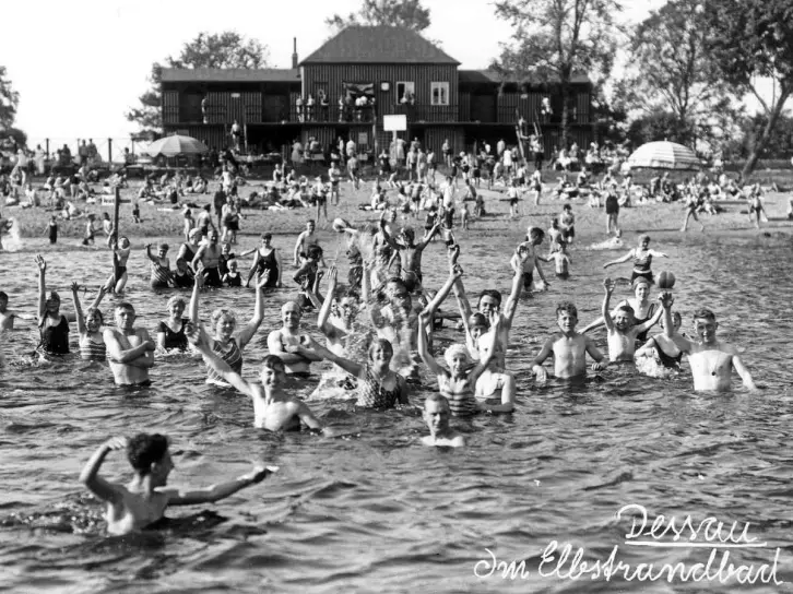 Etwa 100 Menschen in einer Flussbadeanstalt. Vorn steht eine große Gruppe, hebt jubelnd die Arme und schaut zur Kamera. Andere Leute halten sich auf dem Strand auf. Dort steht auch ein Gebäude aus Holz