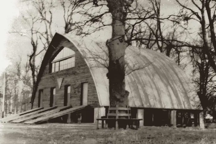 Bootshaus mit Holzfassade, schräg von vorne fotografiert. Im Vordergrund ein Baum mit umlaufender Sitzbank