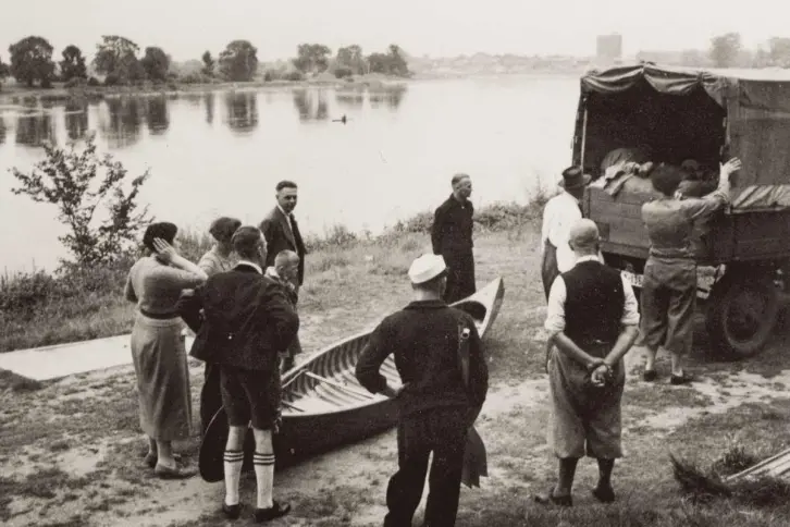 Ein kleiner CanAdier soll auf einen Lkw verladen werden. Etwa zehn Leute warten, dass sie mit der Arbeit beginnen können. Im Hintergrund ist ein Fluss zu sehen. Die Aufnahme stammt aus den früher 30er Jahren.