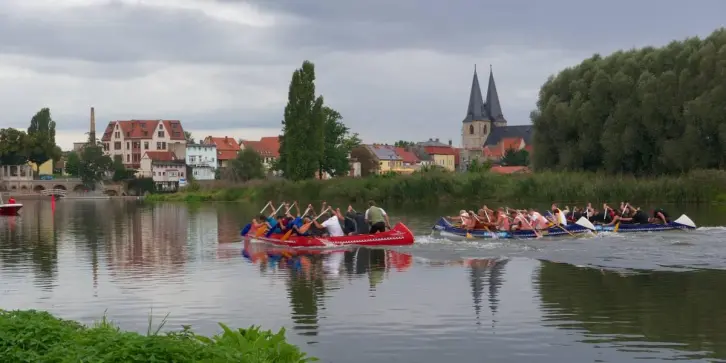 Zwei 10er-Canadier auf einem kleinen Fluss vor einer Stadtsilhouette mit einer Kirche.