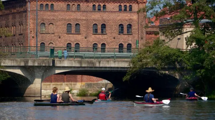Mehrere Kanuten vor einer kleineren Autobrücke und den historischen Mauern einer Großmühle.