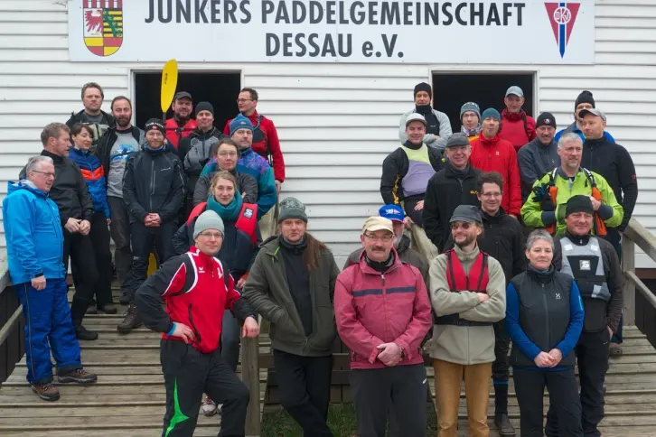 Ein Gruppe von 30 Menschen, viele von ihnen mit Schwimmwesten, haben sich vor einem Bootshaus versammelt. An der Fassade steht Junkers Paddelgemeisnchaft.