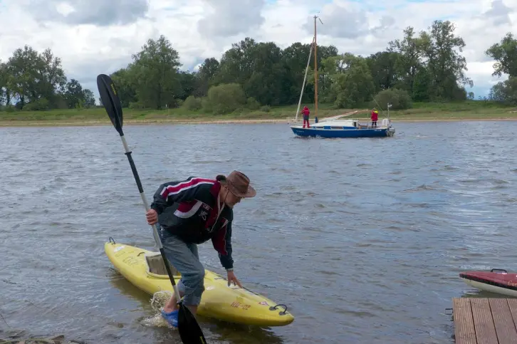 Ein Mann zieht sein Kajak ans Ufer, im Hintergrund ist ein kleines Segelboot unterwegs.