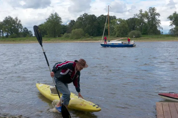 Ein Mann zieht sein Kajak ans Ufer, im Hintergrund ist ein kleines Segelboot unterwegs.