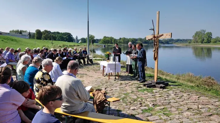 Menschen sitzen im Halbkreis auf Bänken am Flussufer, an dessen Ufern ein improvisiertes Holzkreuz errichtet wurde. Ein Tisch dient als Altar, hinter dem vier Personen stehen.
