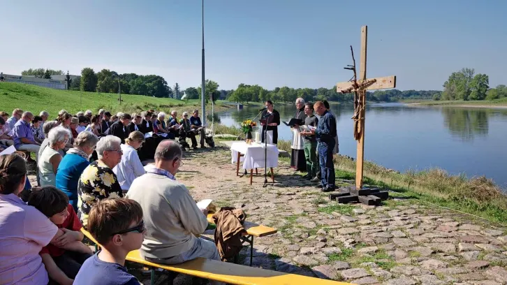 Menschen sitzen im Halbkreis auf B&auml;nken am Flussufer, an dessen Ufern ein improvisiertes Holzkreuz errichtet wurde. Ein Tisch dient als Altar, hinter dem vier Personen stehen.