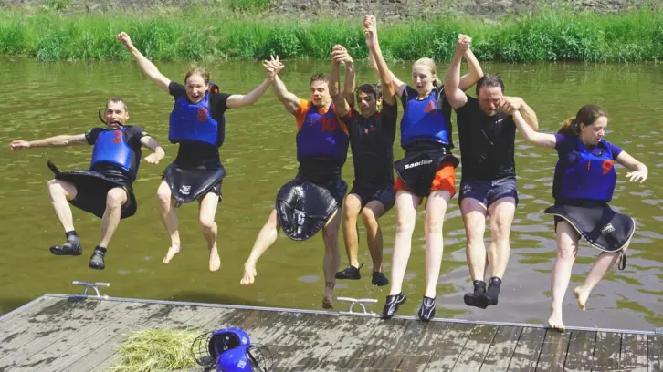 Sieben Personen mit Schwimmwesten springen gemeinsam rückwärts von einem Steg ins Wasser.