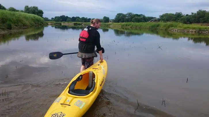 Ein Mann mit blonden Haaren, die zu einem Zopf gebunden sind, steht in flachem Wasser und hält ein gelbes Kajak. Er trägt eine schwarze Jacke, eine kurze Hose und eine rote Schwimmweste. In der rechten Hand hält er ein Doppelpaddel. Die Umgebung ist eine ruhige Flusslandschaft mit flachem, spiegelndem Wasser, wenigen Wasserpflanzen und grünen Ufern. Im Hintergrund ist eine Bogenbrücke zu sehen, umgeben von Bäumen und Wiesen. Der Himmel ist teilweise bewölkt.