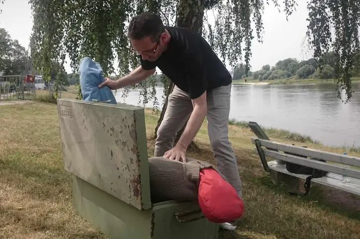 Ein Mann mit Brille, schwarzem T-Shirt und grauer Hose beugt sich über eine offene, grüne Metallkiste und verstaut darin einen menschenähnlichen Dummy. Der Dummy besteht aus einem mit Stoff gefüllten Anzug. Die Szene spielt sich auf einer Wiese unter einem Baum ab, mit Blick auf einen ruhigen Fluss.