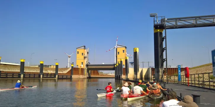 Mehrere Kanuten warten in einer Schleuse, während sich das Schleusentor im Hintergrund befindet. Die Schleusenanlage besteht aus massiven gelben Türmen mit schwarzem Dach und einer zentralen Brücke. Die Schleusenwände sind mit hohen, schwarz-gelben Dalben ausgestattet.