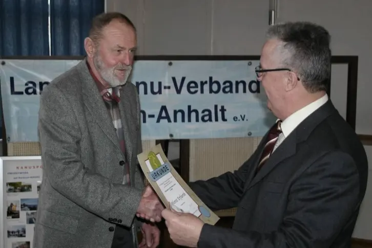 Ein älterer Mann in grauem Sakko erhält eine Urkunde von einem Mann im Anzug. Beide geben sich die Hand. Im Hintergrund hängt ein Banner des Landes-Kanu-Verbands Sachsen-Anhalt e.V.