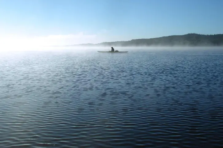 Ein einzelner Kajakfahrer paddelt auf einem ruhigen See, umgeben von morgendlichem Nebel. Der Himmel ist klar und blau, im Hintergrund sind bewaldete Hügel zu sehen. Die Szene strahlt Ruhe und Einsamkeit aus.