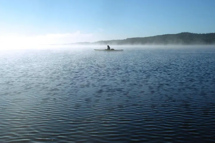 Ein einzelner Kajakfahrer paddelt auf einem ruhigen See, umgeben von morgendlichem Nebel. Der Himmel ist klar und blau, im Hintergrund sind bewaldete H&uuml;gel zu sehen. Die Szene strahlt Ruhe und Einsamkeit aus.