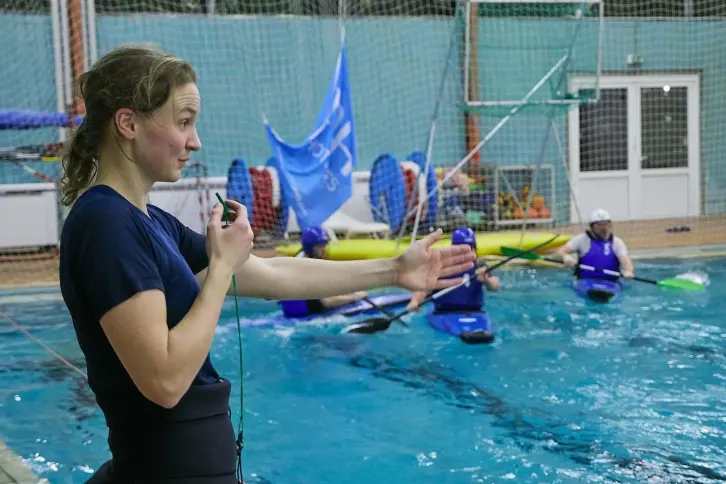 Eine Schiedsrichterin mit Pfeife gibt Anweisungen bei einem Kanupolo-Spiel in einem Hallenbad. Im Wasser spielen mehrere Sportler in blauen Trikots auf Kajaks. Im Hintergrund sind Netze, Ausrüstung und eine blaue Fahne zu sehen.