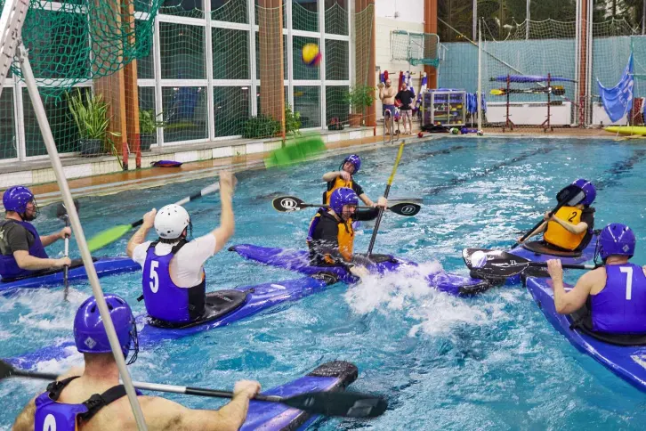 Kanupolo-Spiel in einem Hallenbad: Spieler in blauen und orangefarbenen Trikots kämpfen auf Kajaks um den Ball. Ein Spieler wirft den Ball in Richtung Tor, während andere mit Paddeln eingreifen. Im Hintergrund sind Netze, Pflanzen und Ausrüstung zu sehen.