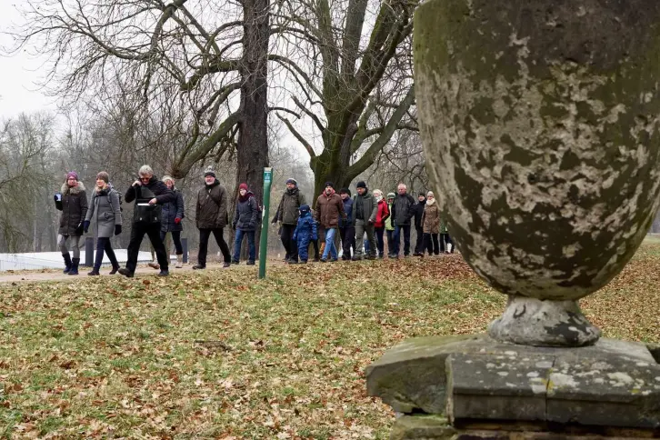 Eine Gruppe von Menschen in warmer Kleidung geht auf einem Parkweg entlang. Die Bäume sind kahl, und der Boden ist mit Herbstlaub bedeckt. Im Vordergrund steht eine verwitterte Steinurne. Der Himmel ist grau.