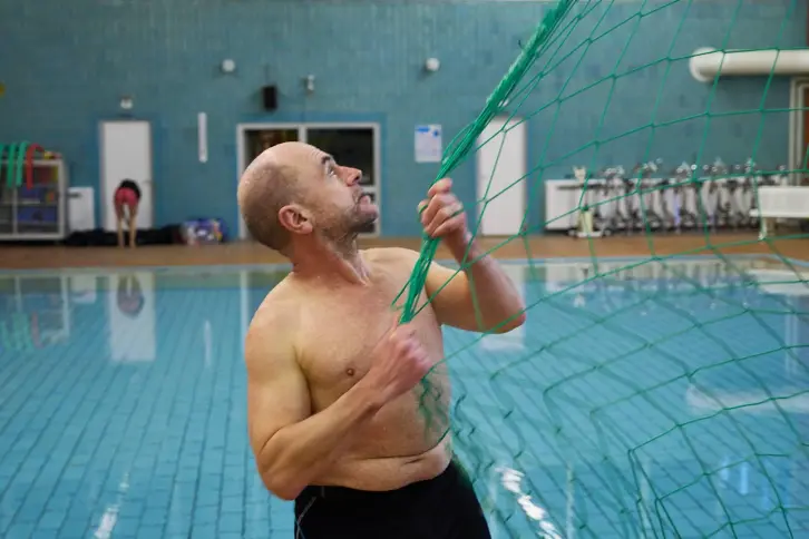 Ein Mann mit nacktem Oberkörper befestigt ein grünes Netz an einem Tor in einem Hallenbad. Er schaut konzentriert nach oben. Im Hintergrund sind das Schwimmbecken, eine Person in Sportkleidung und verschiedene Ausrüstungsgegenstände zu sehen.