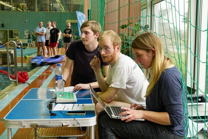 Drei Personen sitzen an einem Tisch am Beckenrand eines Hallenbads und betreuen ein Kanupolo-Turnier. Sie arbeiten mit Laptop, Uhr und Notizen. Im Hintergrund stehen Spieler und Boote am Wasser, ein grünes Netz trennt das Spielfeld vom Zuschauerbereich.