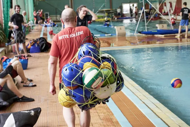 Ein Mann mit rotem T-Shirt und der Aufschrift „BITTE FOLGEN!“ trägt ein Netz voller bunter Bälle und Helme am Beckenrand eines Hallenbads. Um ihn herum sitzen und stehen weitere Personen, im Wasser sind Kanupolo-Boote zu sehen.