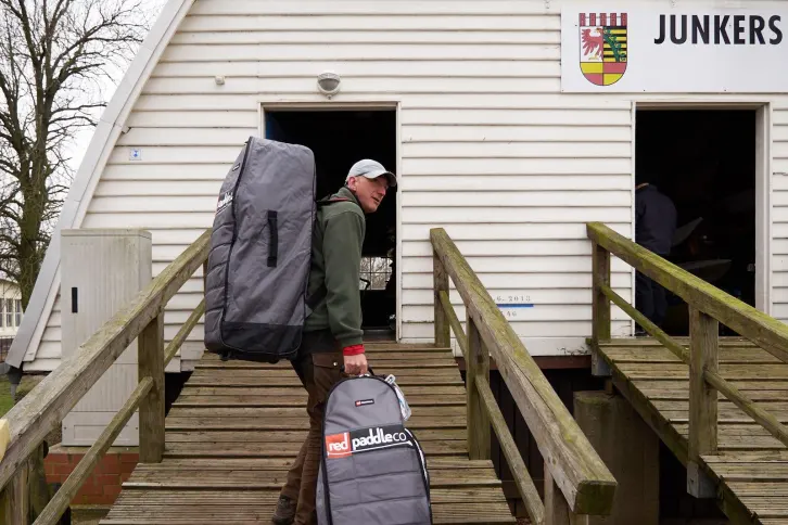 Ein Mann tr&auml;gt zwei gro&szlig;e Taschen mit SUP-Ausr&uuml;stung eine h&ouml;lzerne Rampe zu einem wei&szlig;en Geb&auml;ude mit dem Schild &bdquo;JUNKERS&ldquo; hinauf. Eine T&uuml;r steht offen, im Inneren sind Boote zu erkennen.