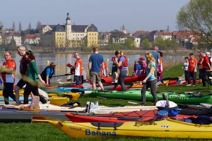 Viele Kajaks am Ufer eines Flusses, dazwischen Paddler, im Hintergrund ein Schloss