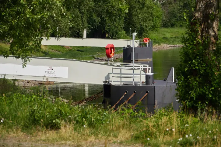 Zwei Anleger für Flusskreuzfahrtschiffe liegen am Ufer eines Flusses. Auf einem ist das Logo der Reederei „Viking“ zu sehen. Die Umgebung ist grün und von Bäumen gesäumt.
