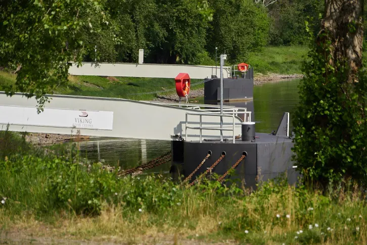 Zwei Anleger f&uuml;r Flusskreuzfahrtschiffe liegen am Ufer eines Flusses. Auf einem ist das Logo der Reederei &bdquo;Viking&ldquo; zu sehen. Die Umgebung ist gr&uuml;n und von B&auml;umen ges&auml;umt.