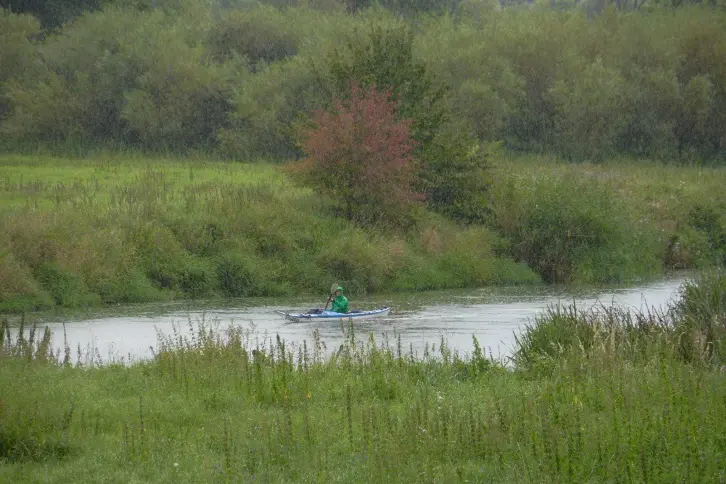 Paddler auf einem kleinen Fluss, der durch eine grüne Landschaft führt. Es regenet etwas.