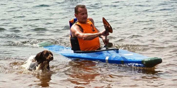 Ein Mann paddelt im Kajak, neben ihm l&auml;uft ein Hund durchs Wasser.
