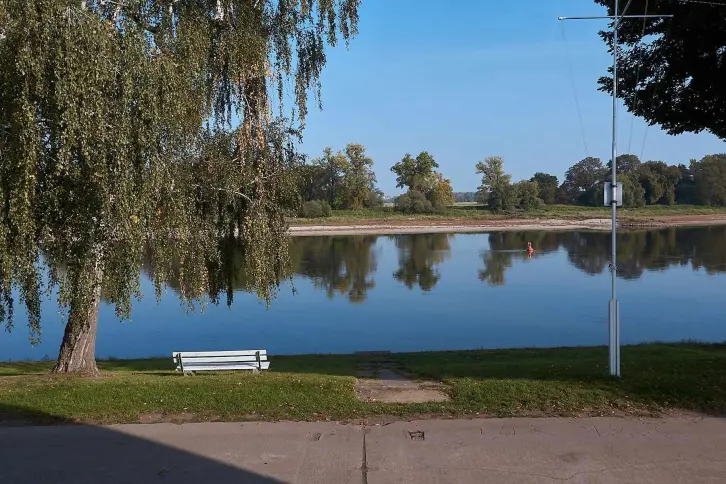 Blick über die Elbe: im Wasser spiegeln sich die Bäume, am diesseitigen Ufer steht eine Bank.