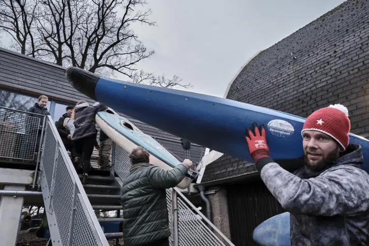 Ein Mann trägt ein Paddelboot auf der Schulter. Andere Personen tragen hinter ihm ein Boot eine Treppe hinaus.