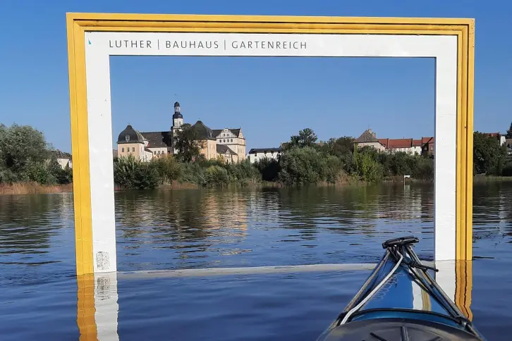 Bei Hochwasser steht ein großer hölzener Rahmen im Wasser, der einem Bilderrahmen ähnelt. Durch diesen sieht man das Schloss Coswig.