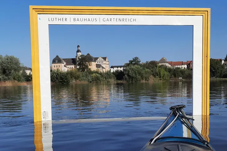 Bei Hochwasser steht ein großer hölzener Rahmen im Wasser, der einem Bilderrahmen ähnelt. Durch diesen sieht man das Schloss Coswig.