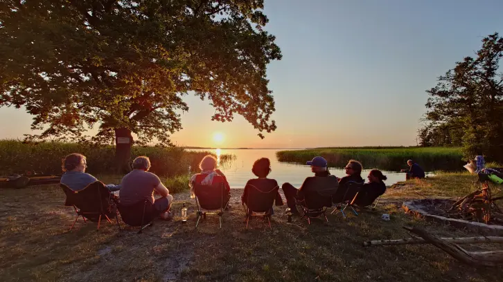 Sechs Menschen sitzen am Wasser und betrachten den Sonnenuntergang.